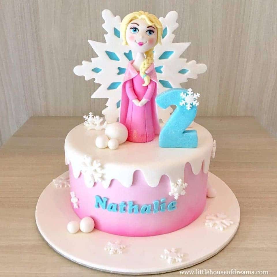 A Frozen themed cake that combines pink, white and blue decoration, and topped with edible Queen Elsa figurine. Little House of Dreams.Source